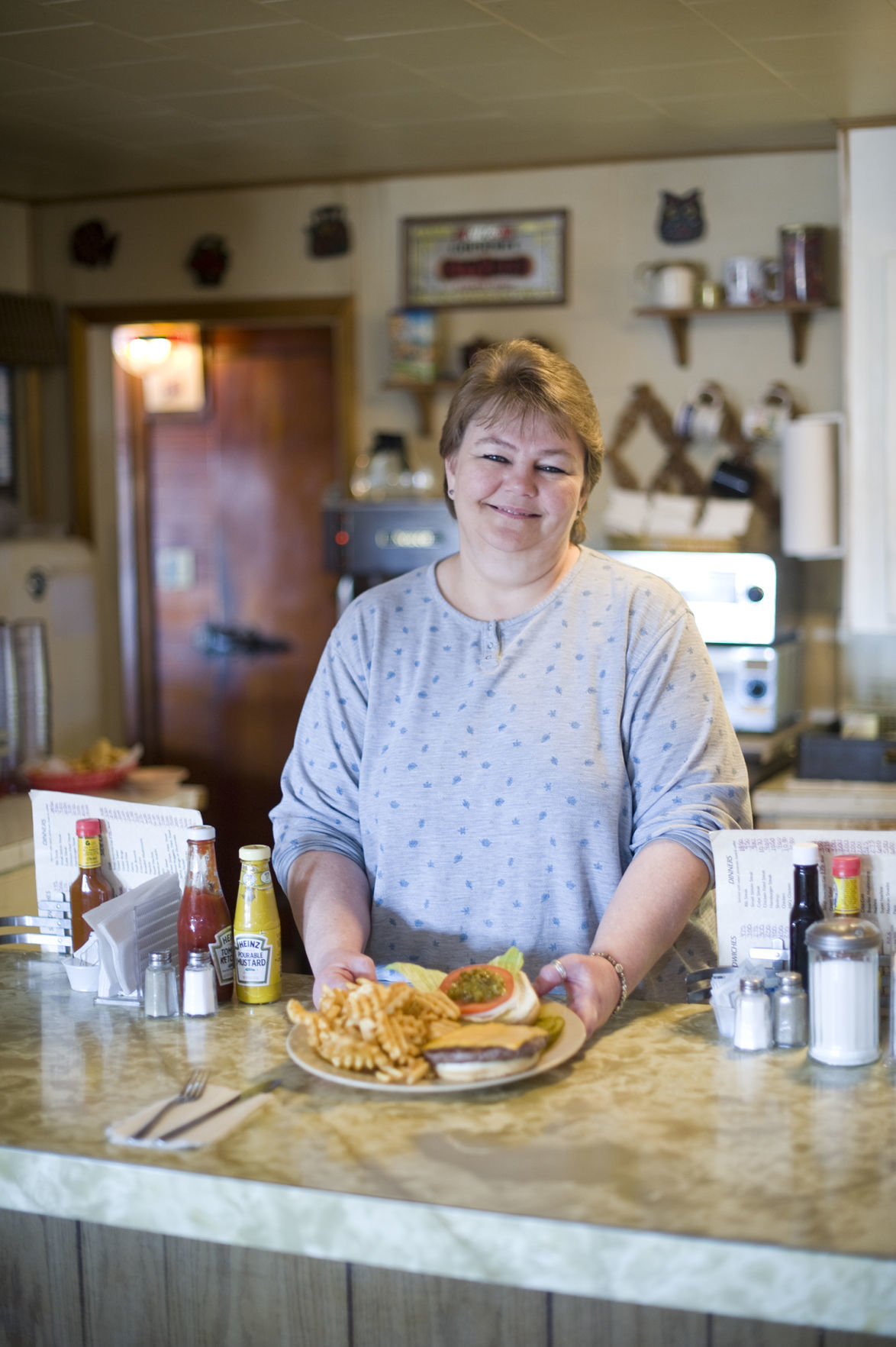 Kim Cook, whose parents Fritzi and Larry Debeau owned Park Grove Cafe before her, pictured at the counter holding Fritzi's Dam Burger with Crisscut fries. Kim's daughters Shandy Stingley and Ali Cook also work at the family business.